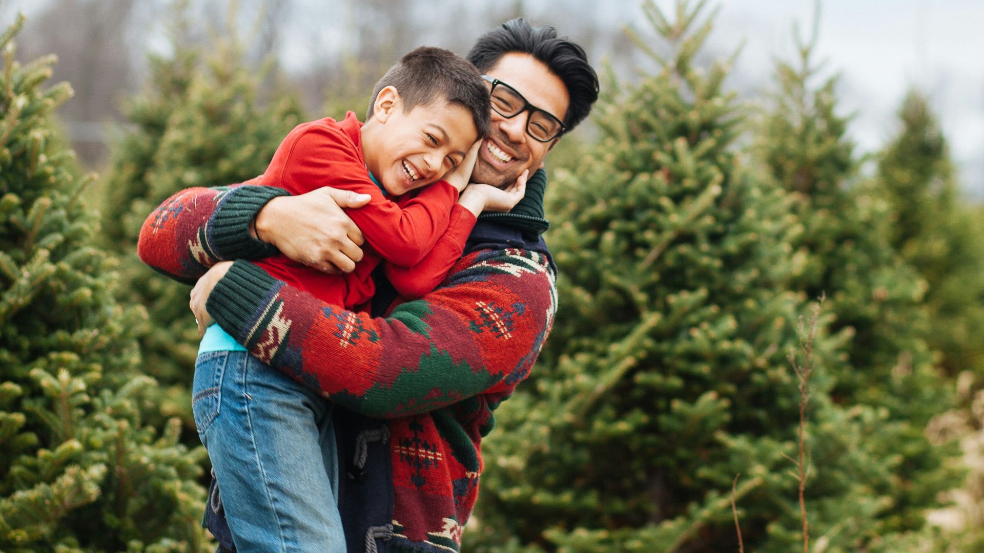 man carrying boy while standing and smiling near pine trees