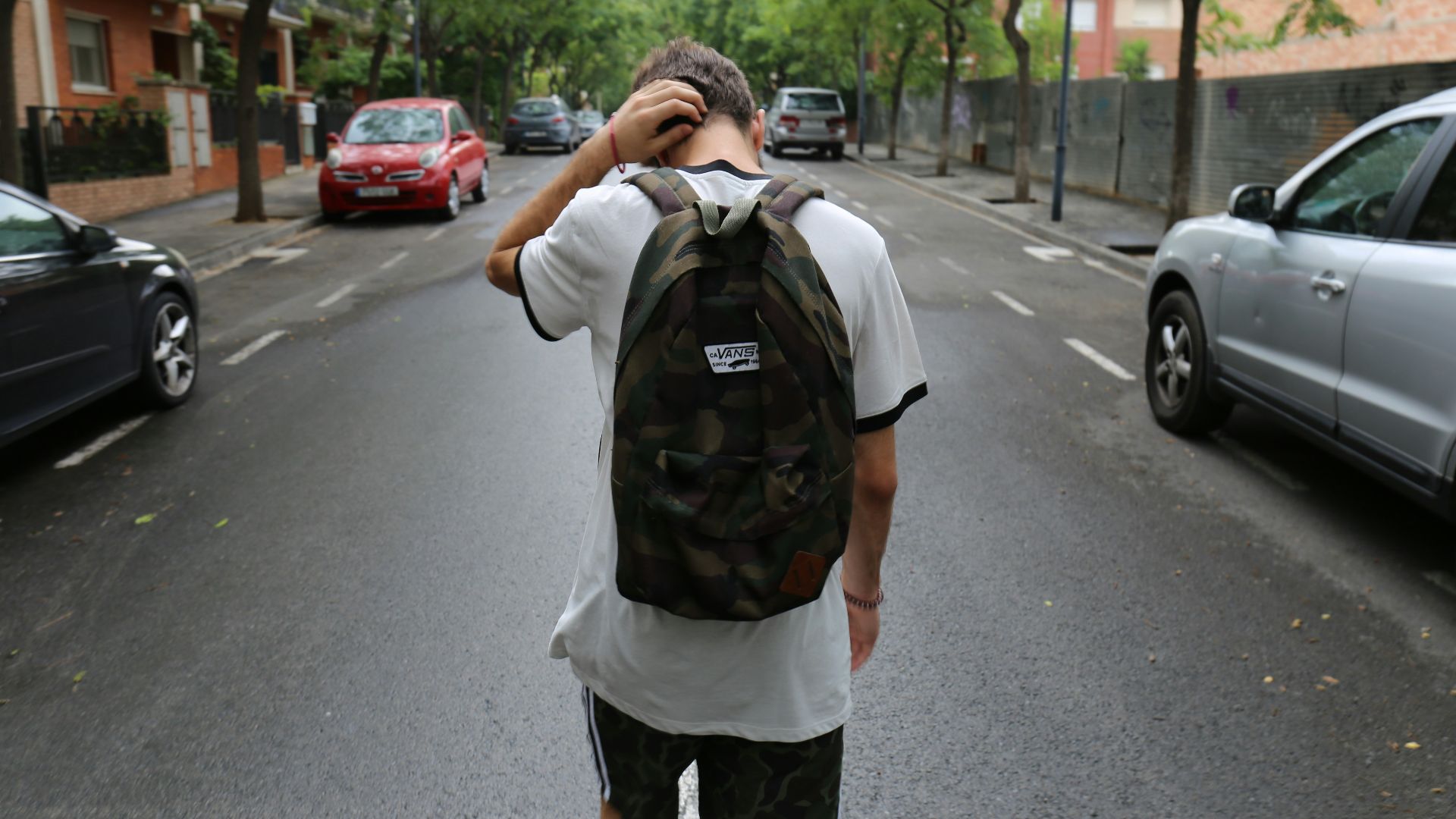 boy wearing white shirt and black shorts carrying backpack standing on black concrete road between vehicles and trees during daytime