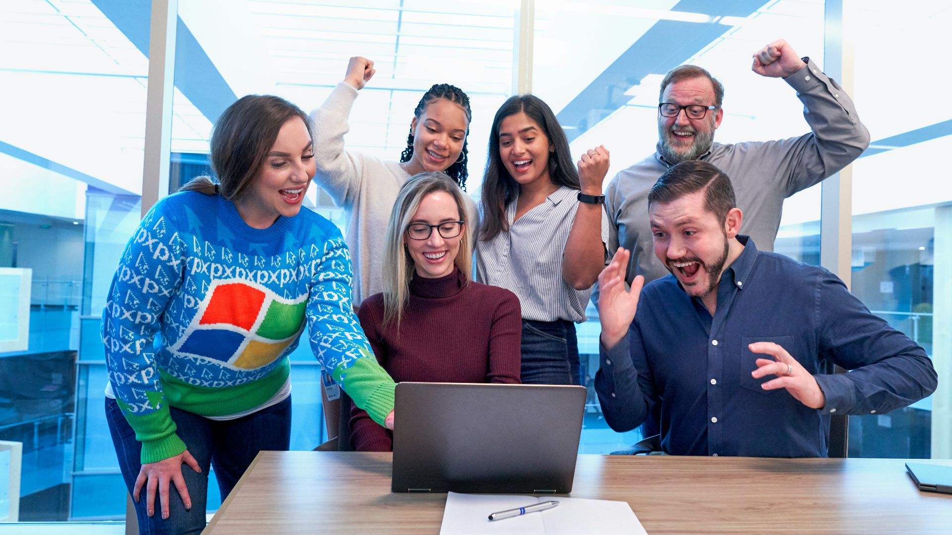 men and women sitting and standing by the table looking happy while staring at laptop