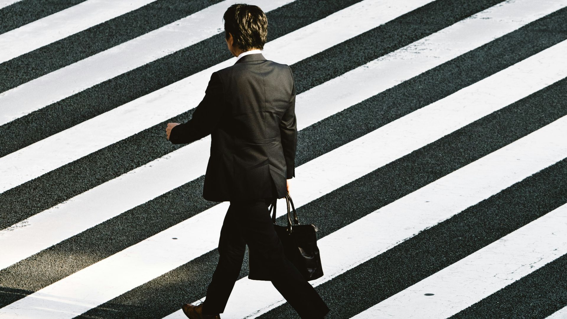 man in black formal suit jacket and pants carrying black bag while walking on pedestrian lane during daytime