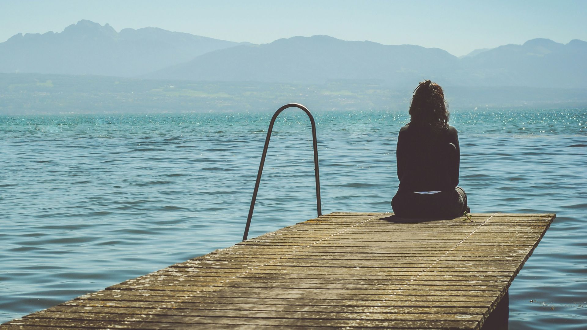 a woman sits on the end of a dock during daytime staring across a lake