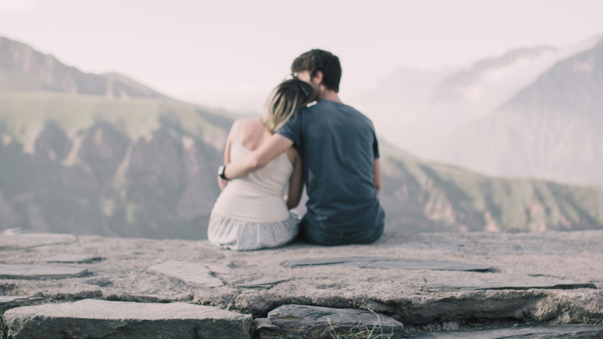 man and woman facing backward while woman's head on man's shoulder on tip of hill