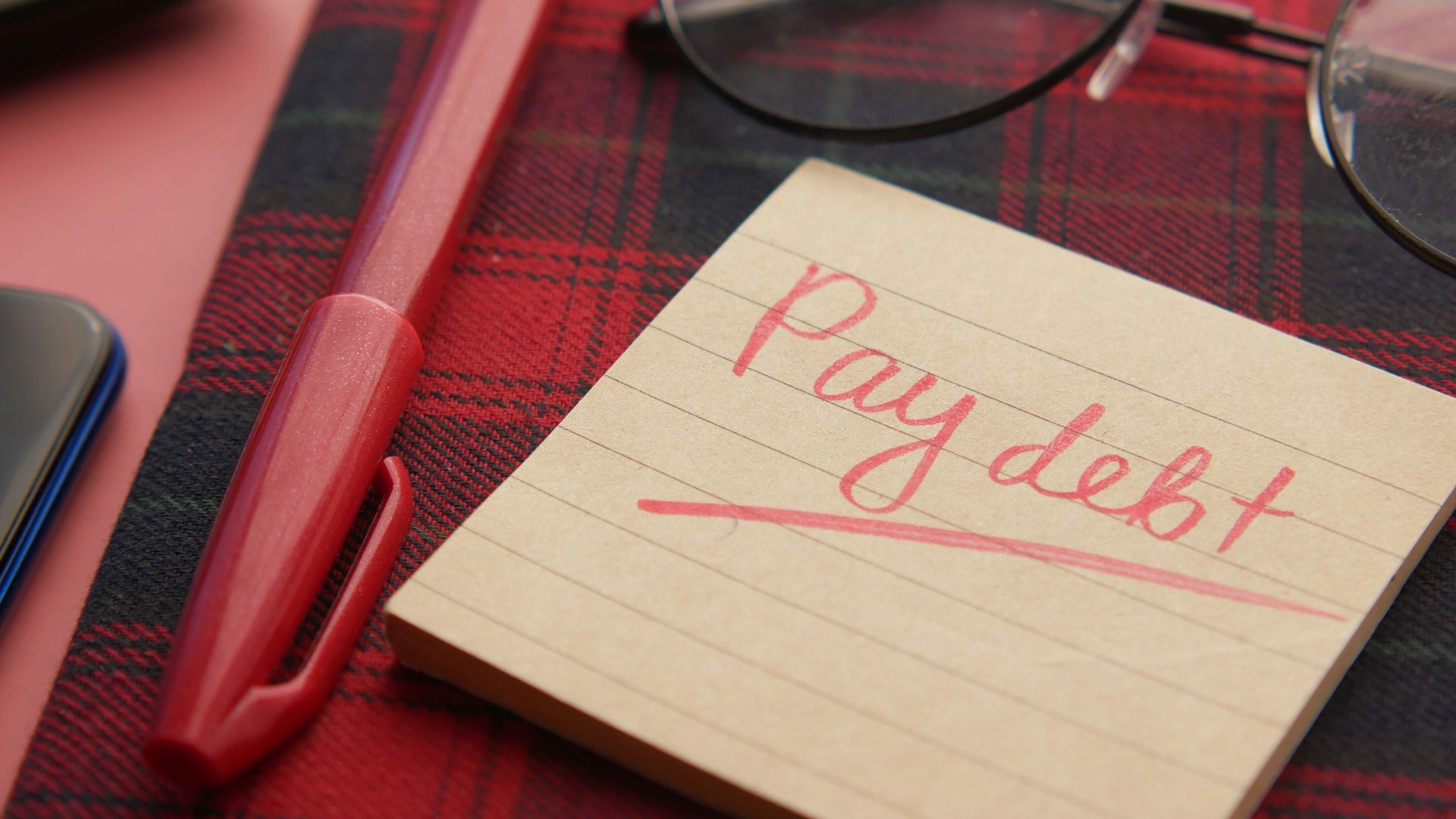a note that says pay debt next to a pen and glasses
