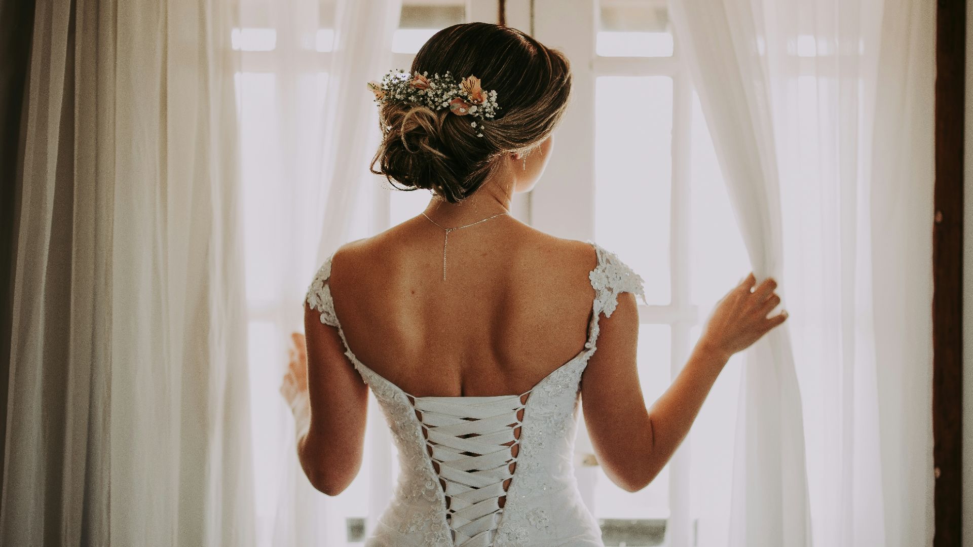 woman in wedding dress standing near window looking outside while holding the curtains