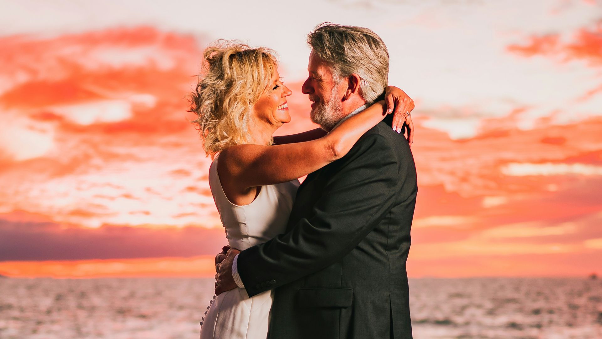 a bride and groom embracing on the beach at sunset