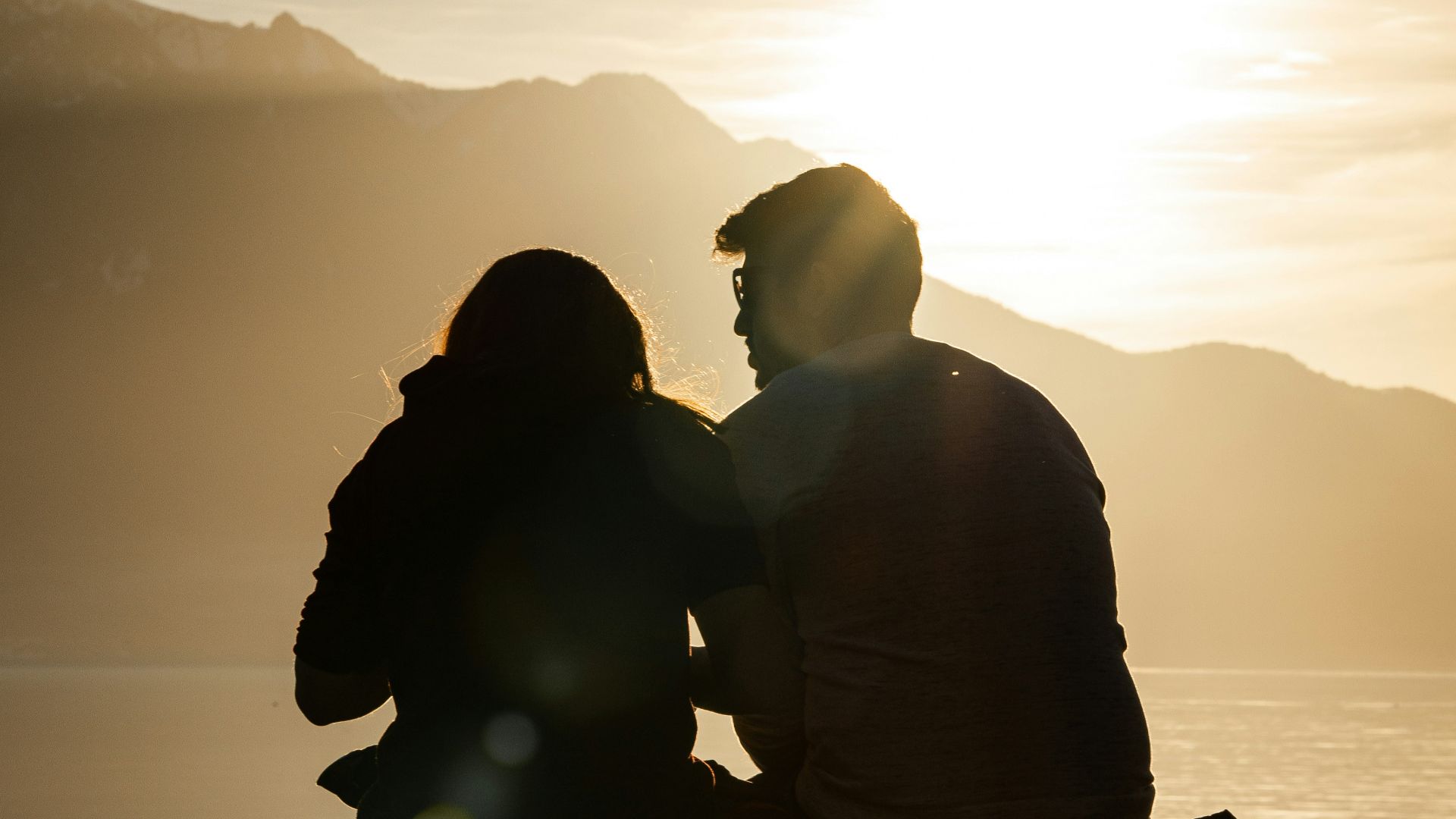 silhouette of man and woman sitting on bench near body of water during sunset