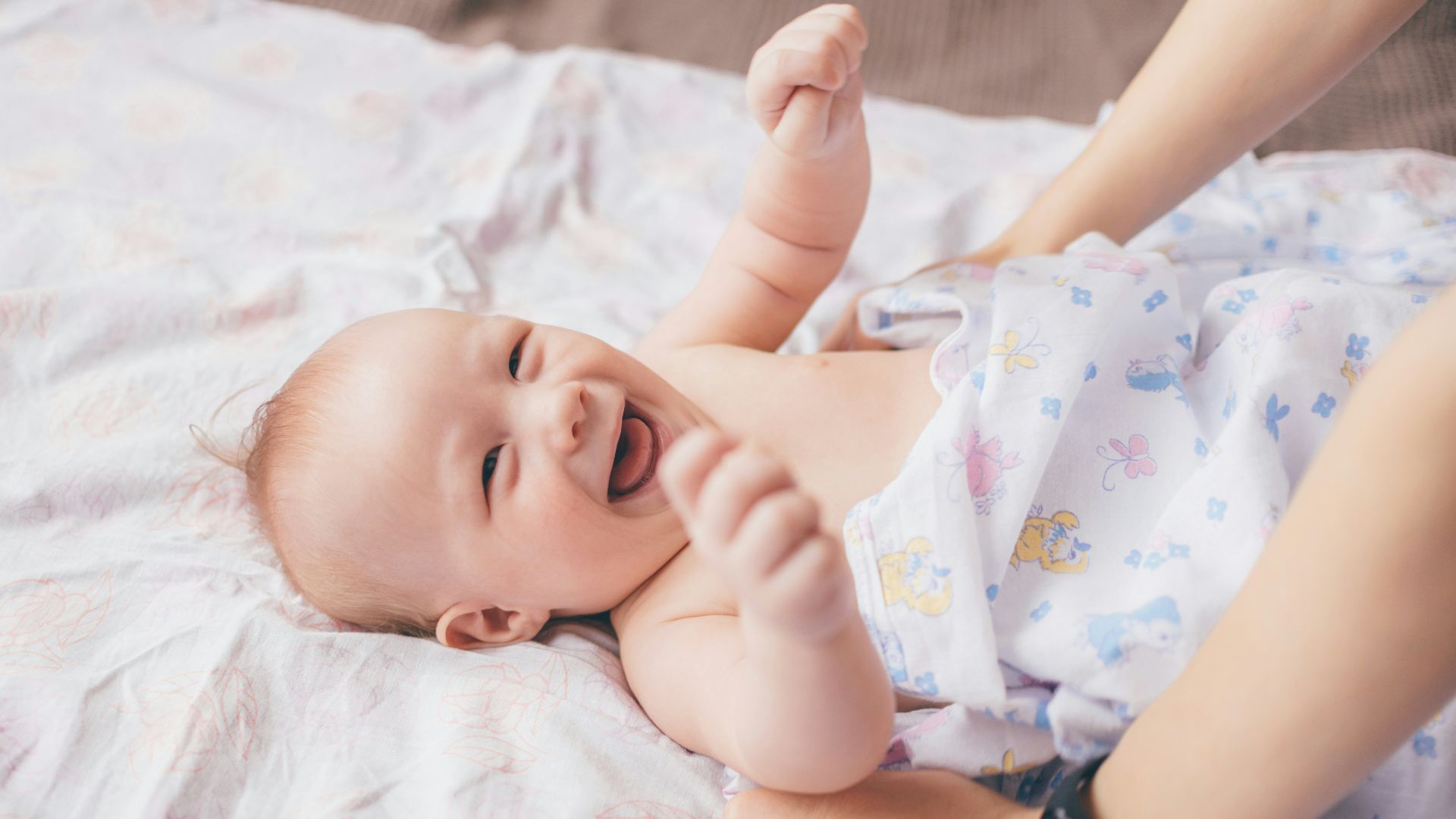 a smiling baby laying on a bed with a woman's arm