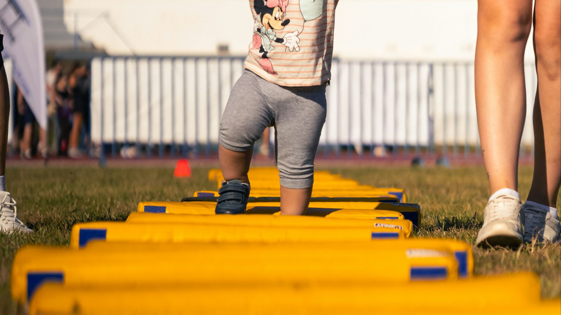 Two women help a toddler walk across yellow hurdles