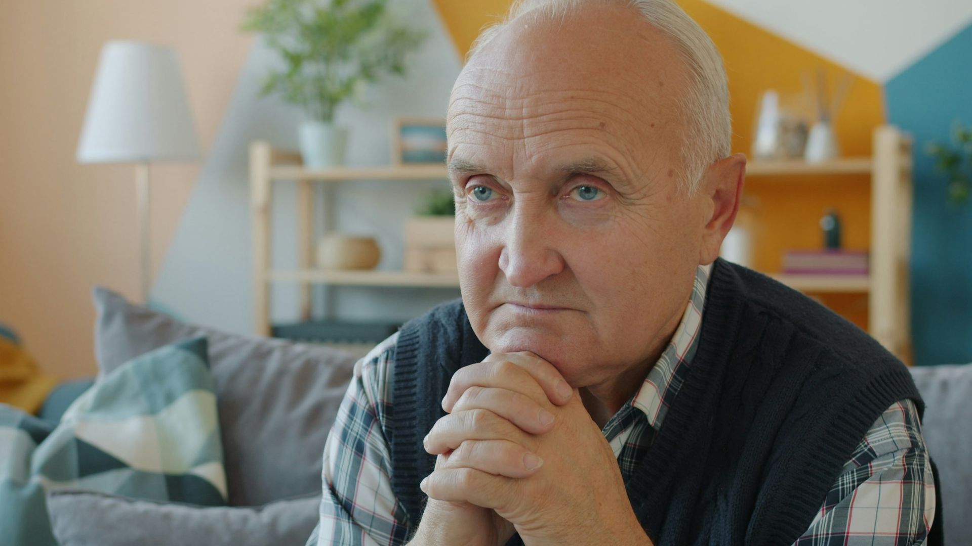 Elderly man sitting on a couch with hands clasped.