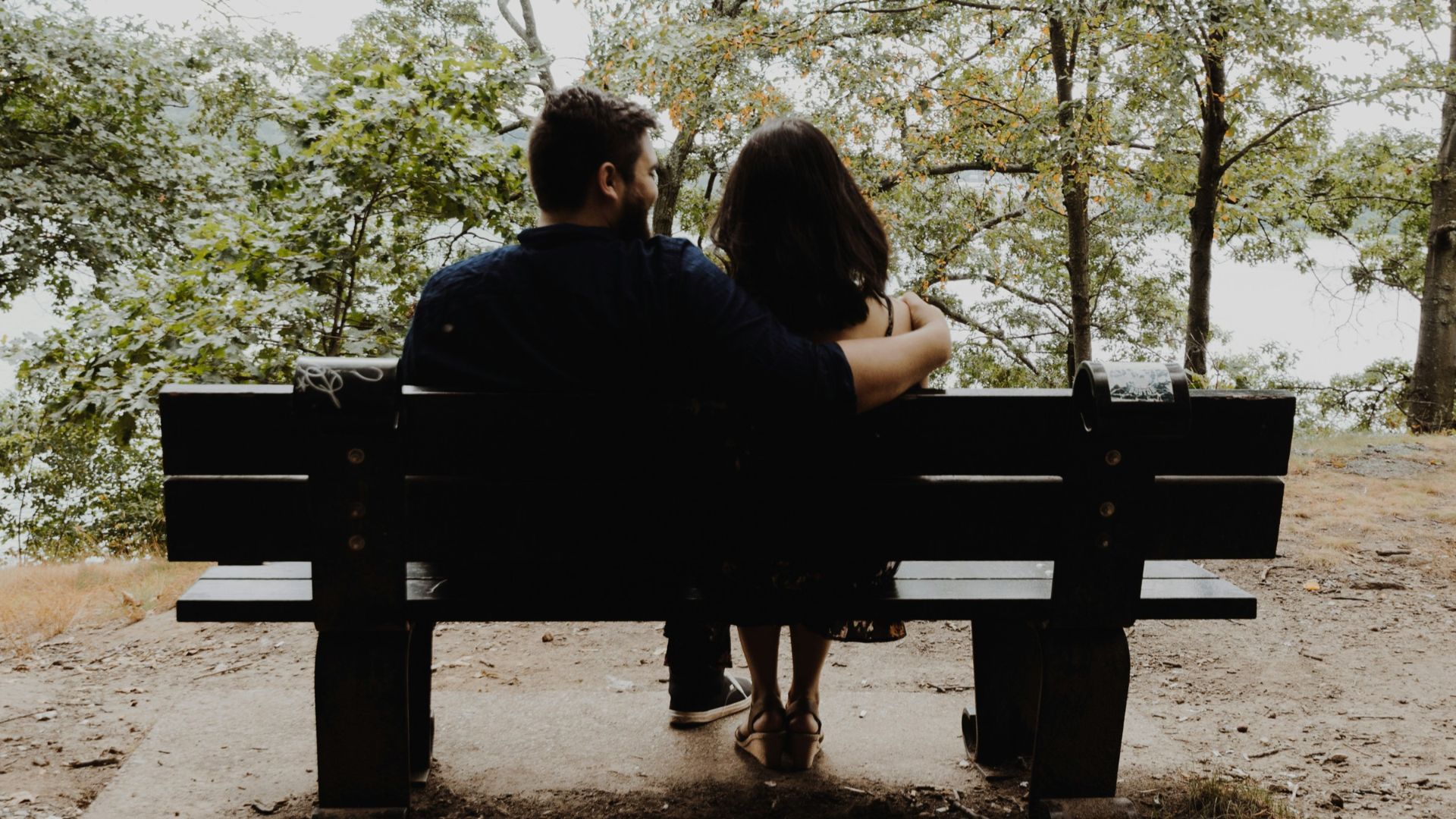 man looking to woman sitting on black wooden bench in front of tall trees during daytime