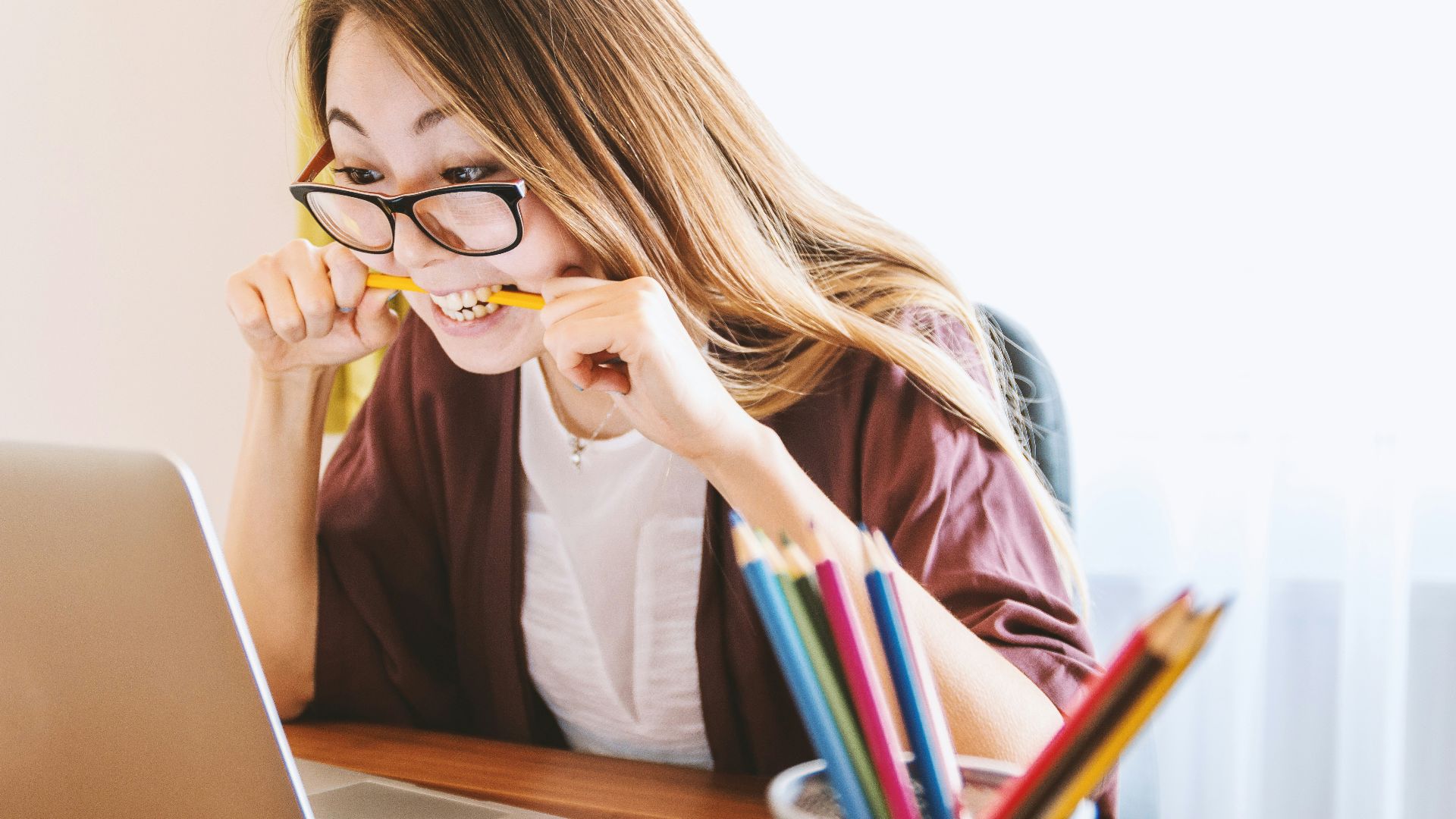 woman biting pencil while sitting on chair in front of computer during daytime