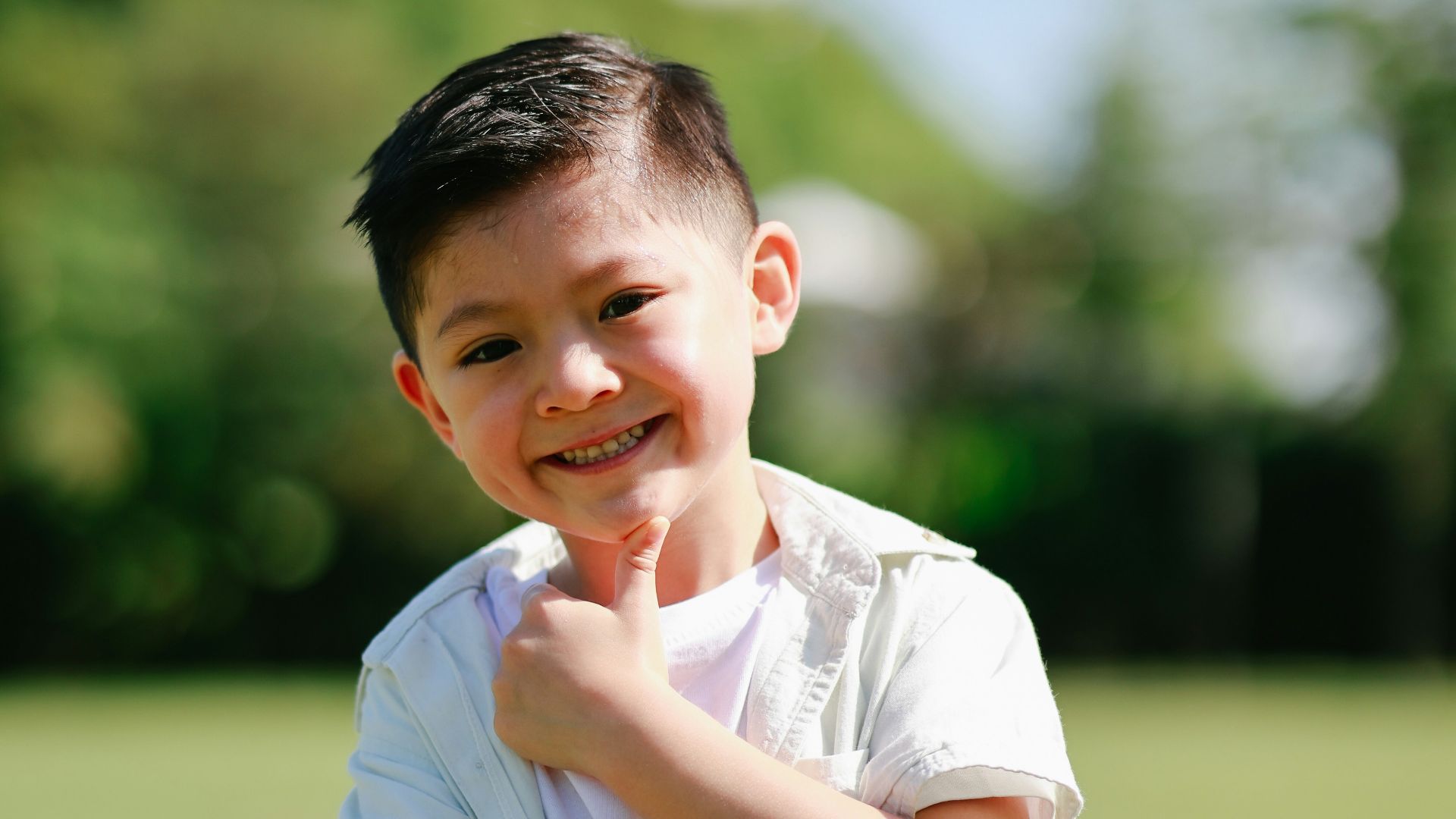 A smiling young boy stands outdoors in a park.