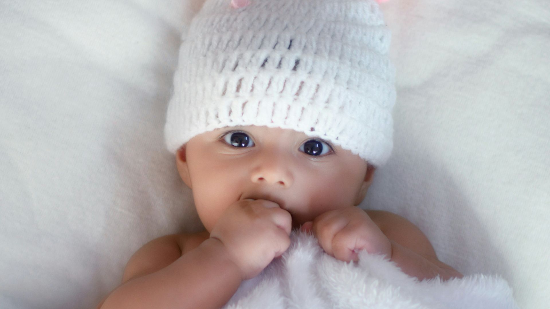 baby in white knit cap lying on white textile