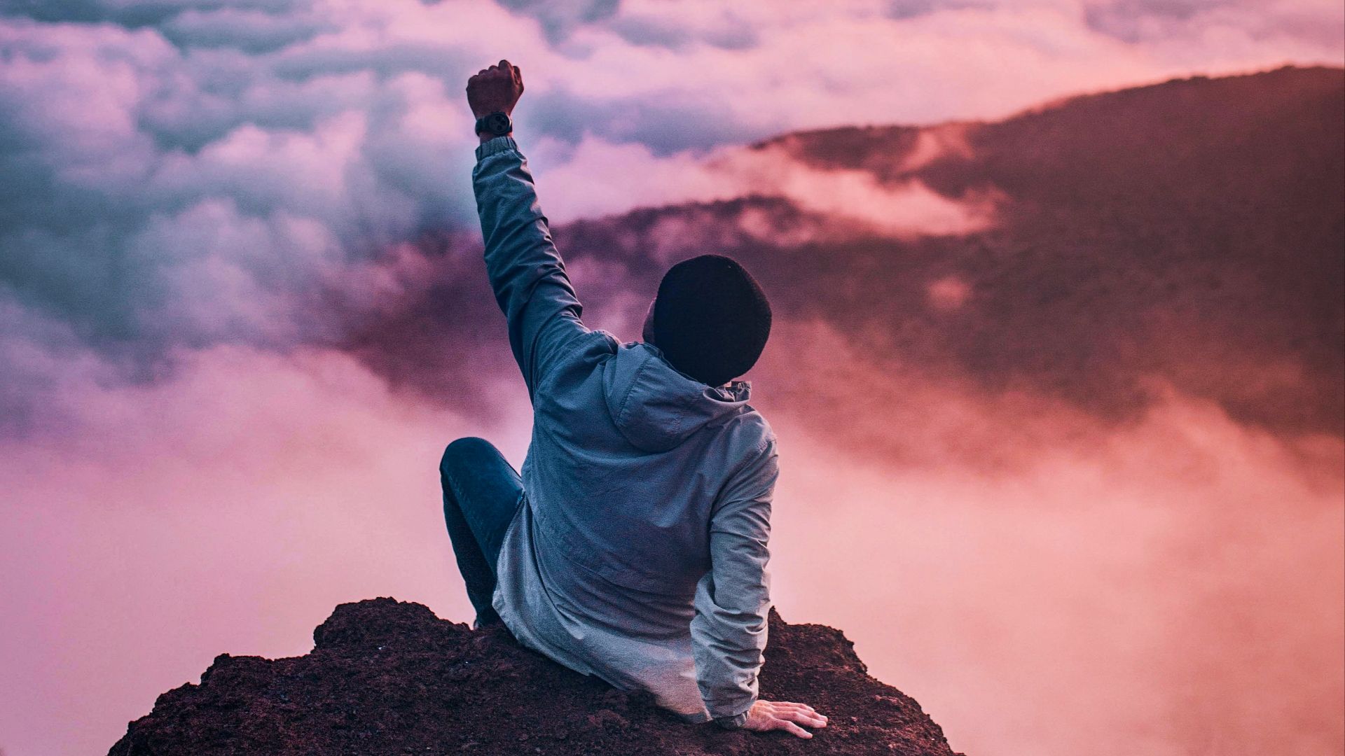 man sitting on mountain cliff facing white clouds rising one hand at golden hour