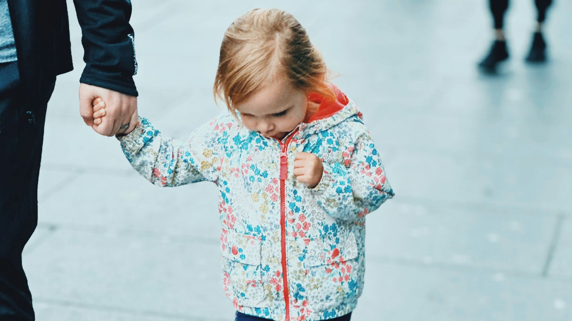 A young girl holds a hand while walking on pavement.