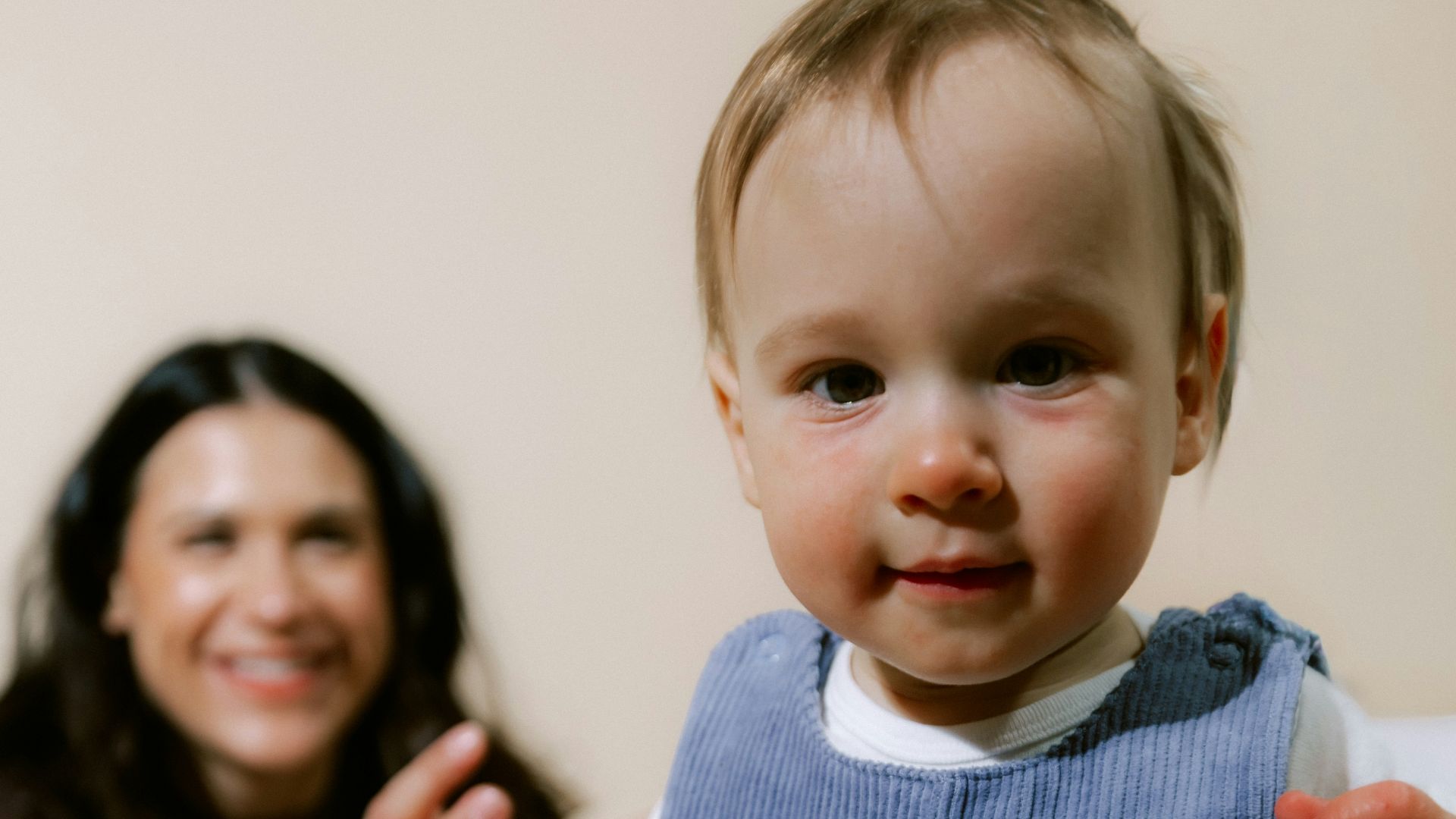 A baby in blue overalls with a woman behind.