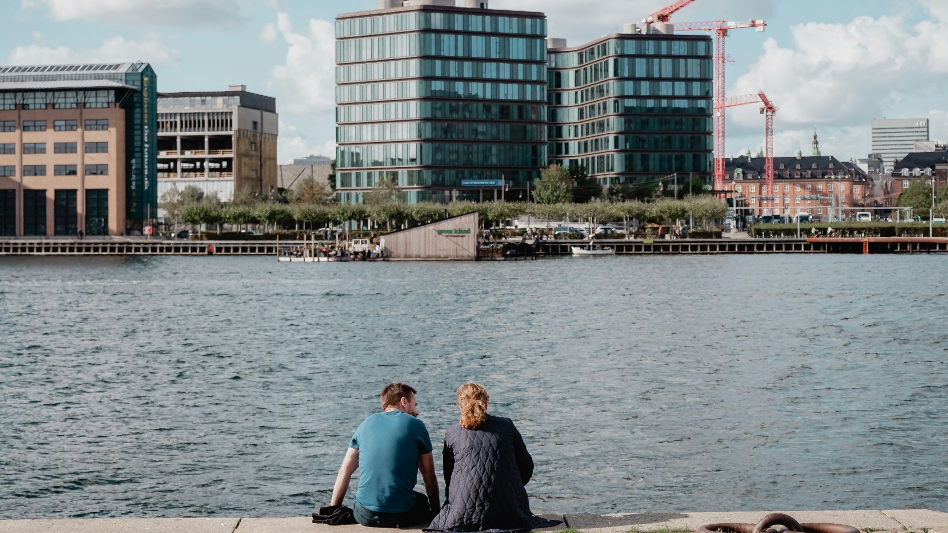man wearing teal crew-neck shirt sitting with woman wearing gray jacket looking at lake