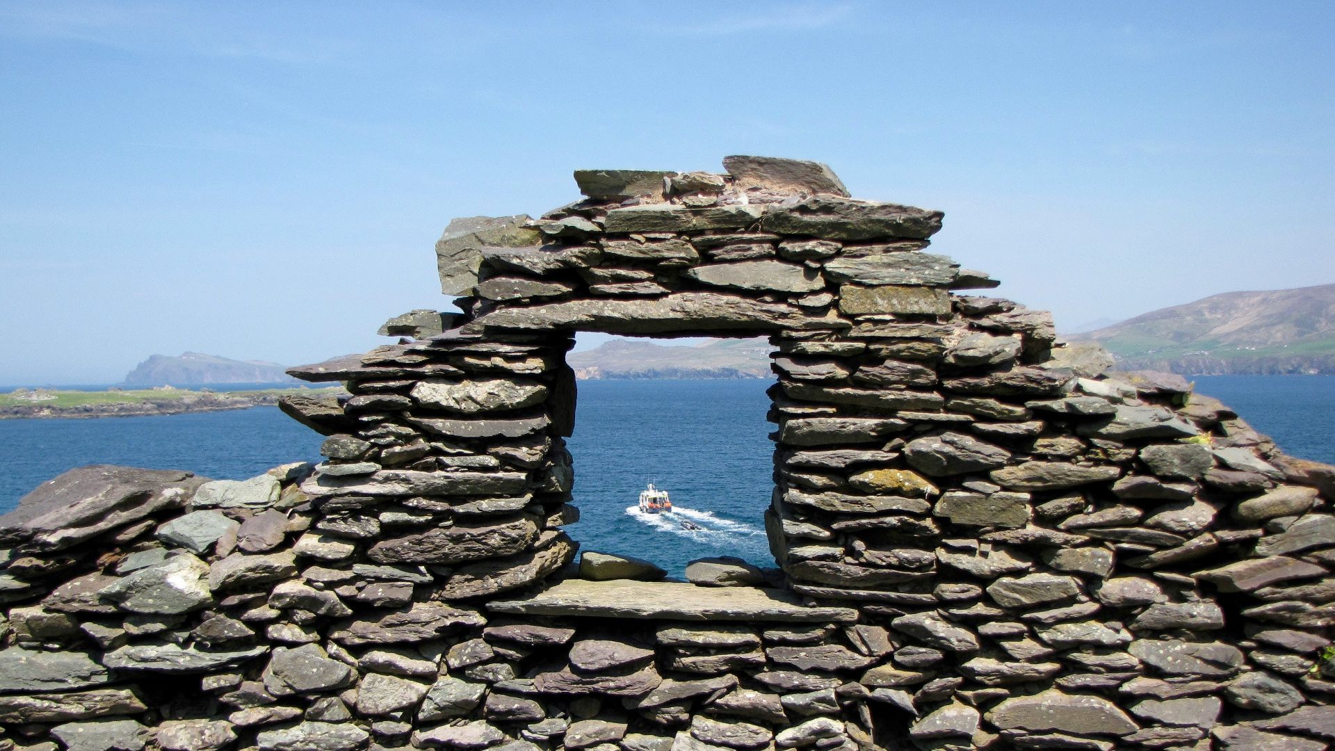 Stone ruins frame a boat on the blue sea.