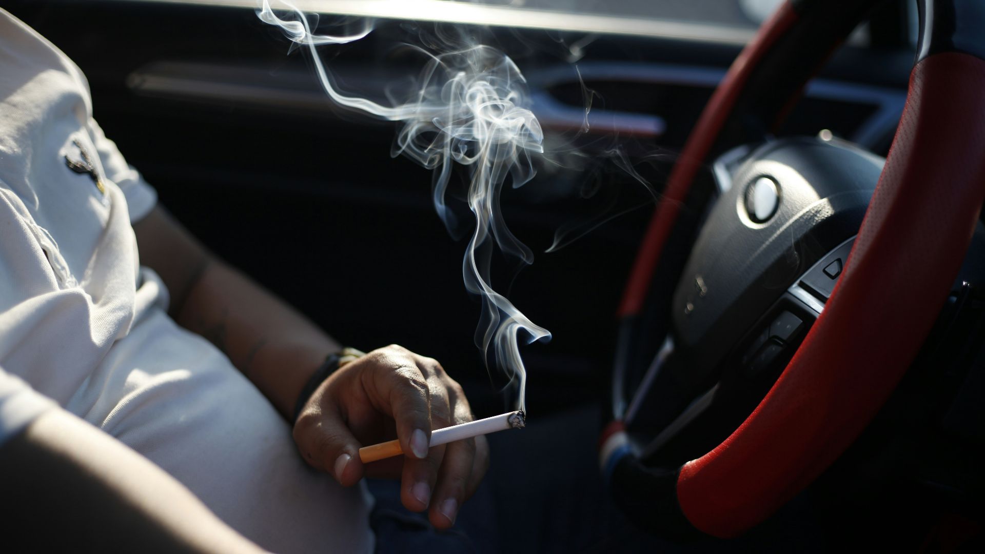 person holding cigarette stick in front of car steering wheel